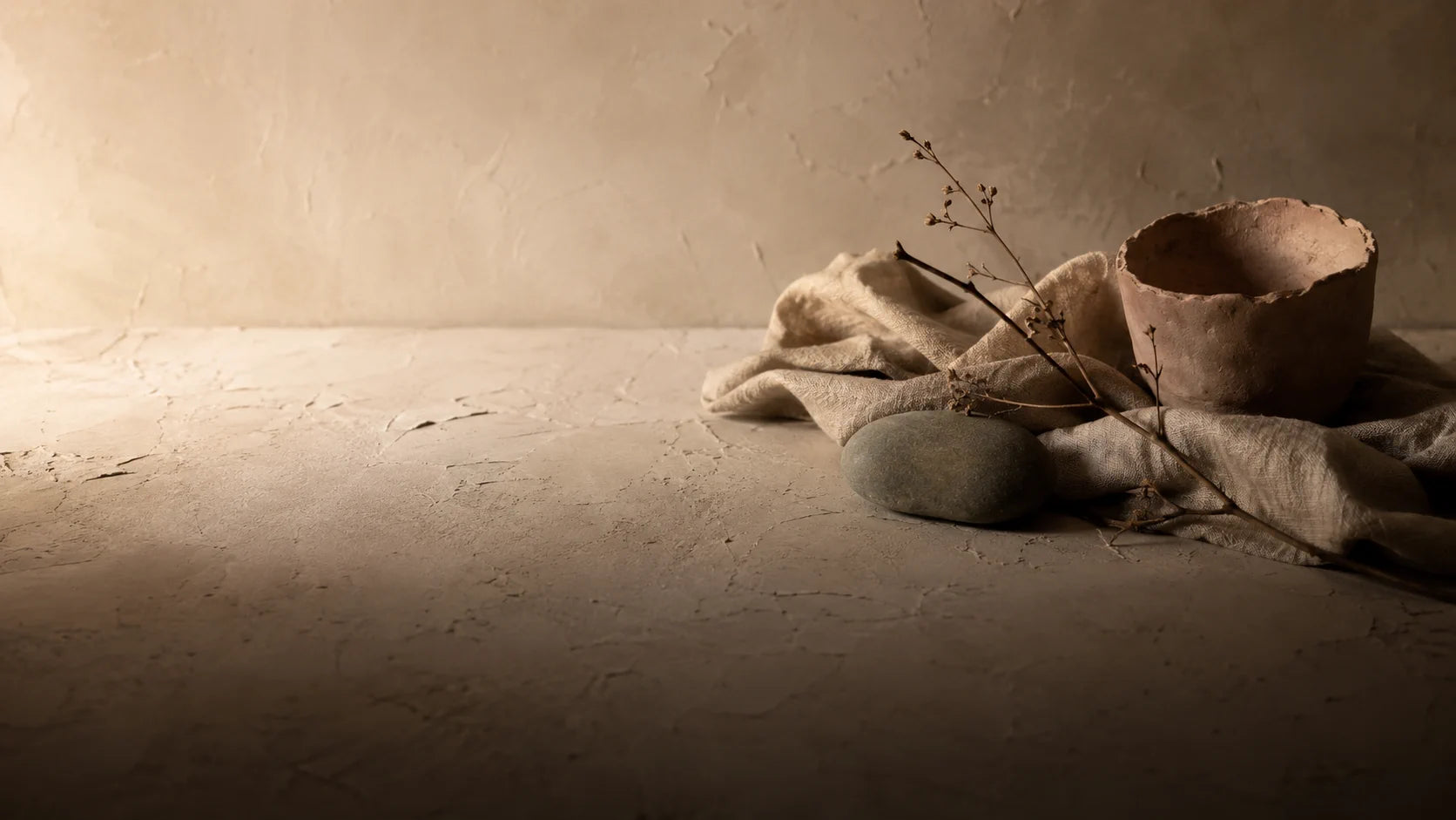 Bodegón wabi-sabi: cuenco de barro sin esmaltar, lino, piedra de río y rama seca sobre muro de yeso
